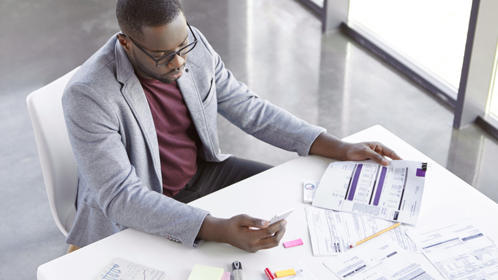 Portrait of successful prosperus dark skinned male works with documents, searches important information on websites, uses modern mobile phone and free internet connection in office, types text