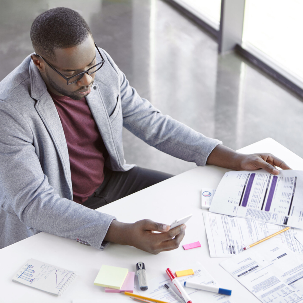 Portrait of successful prosperus dark skinned male works with documents, searches important information on websites, uses modern mobile phone and free internet connection in office, types text